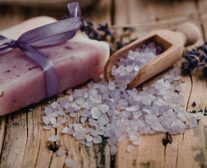 Savon artisanal violet avec un nœud, des sels de bain et de la lavande posés sur un plateau en bois rustique